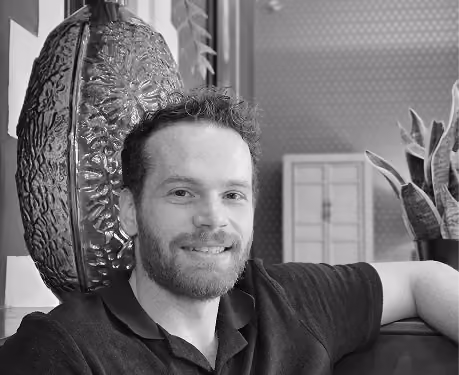 A bearded man with curly hair wearing a dark polo shirt is sitting and smiling indoors near a large decorative vase and plants.