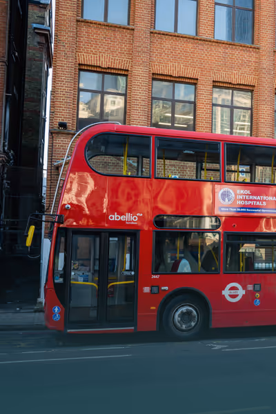 Klasik kırmızı çift katlı Londra otobüsü sokakta park halinde, arka planında tuğla bina ve pencere yansımaları.