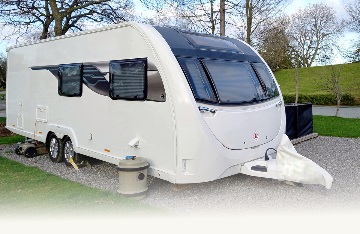 White caravan trailer parked on gravel with closed windows and surrounded by grass and trees.