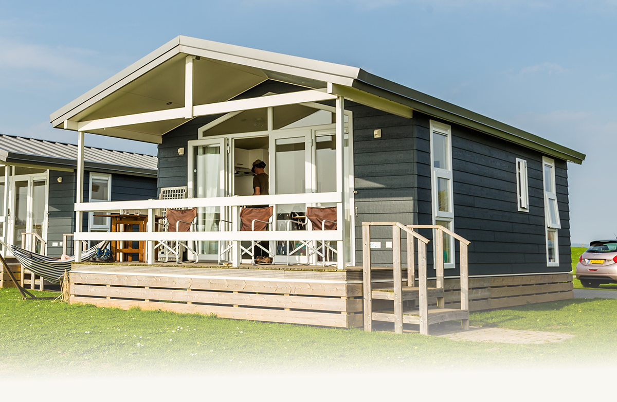 Gray modern cabin with porch, outdoor chairs, and a person inside, set on green grass under blue sky.