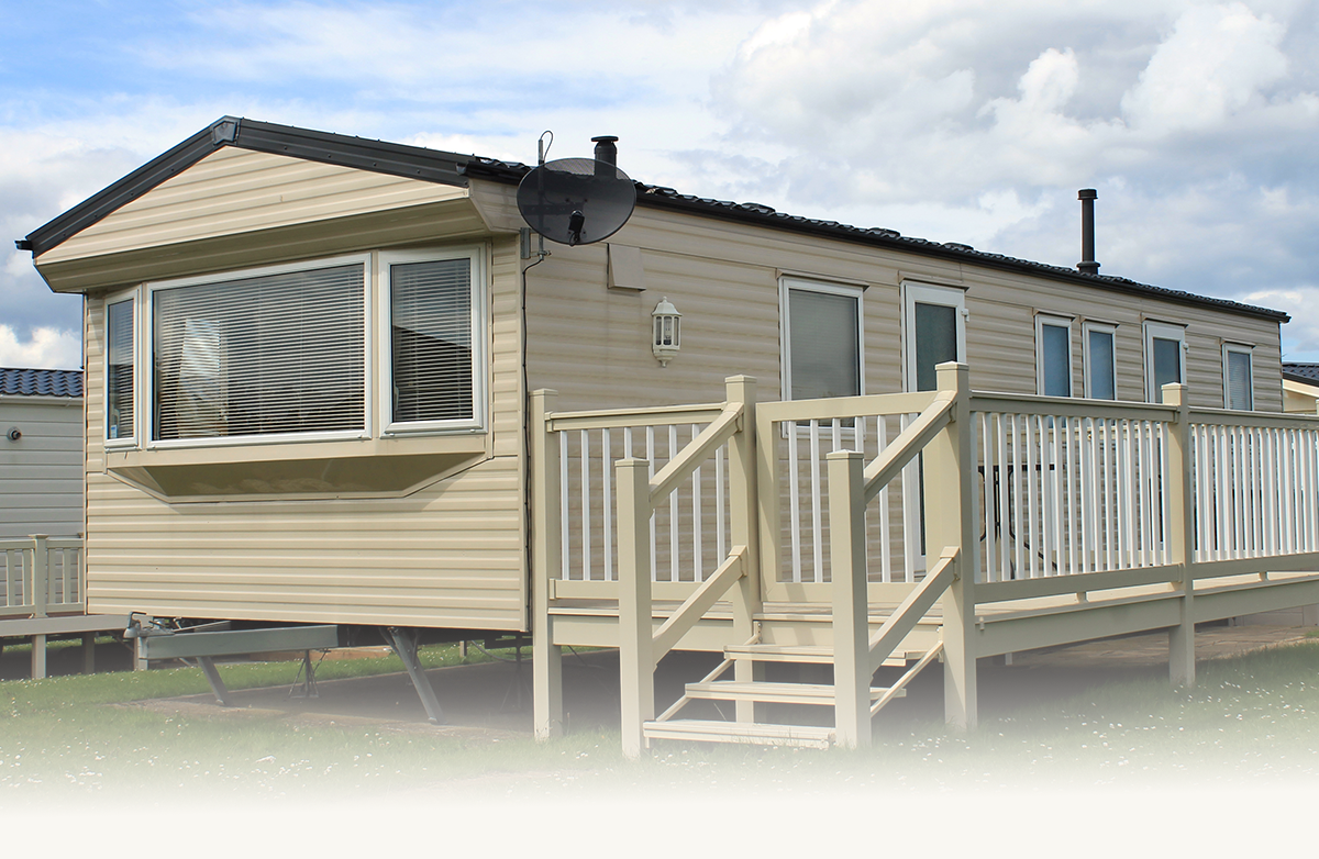 Beige mobile home with large windows, porch with railing, stairs, and satellite dish under cloudy sky.