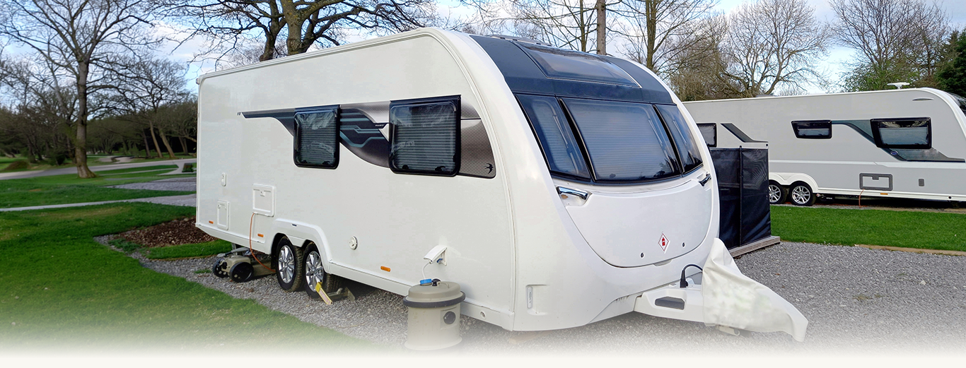 White caravan with black windows parked on gravel near grass and leafless trees.