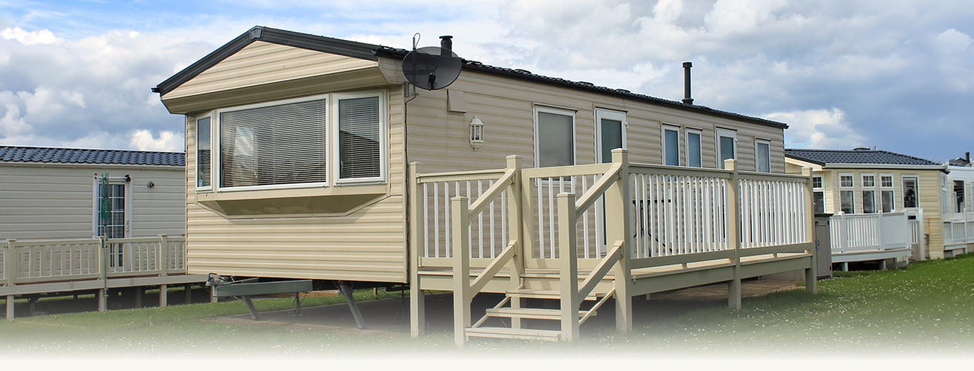 Beige static caravan with white railings and stairs on a grassy area under a cloudy sky.