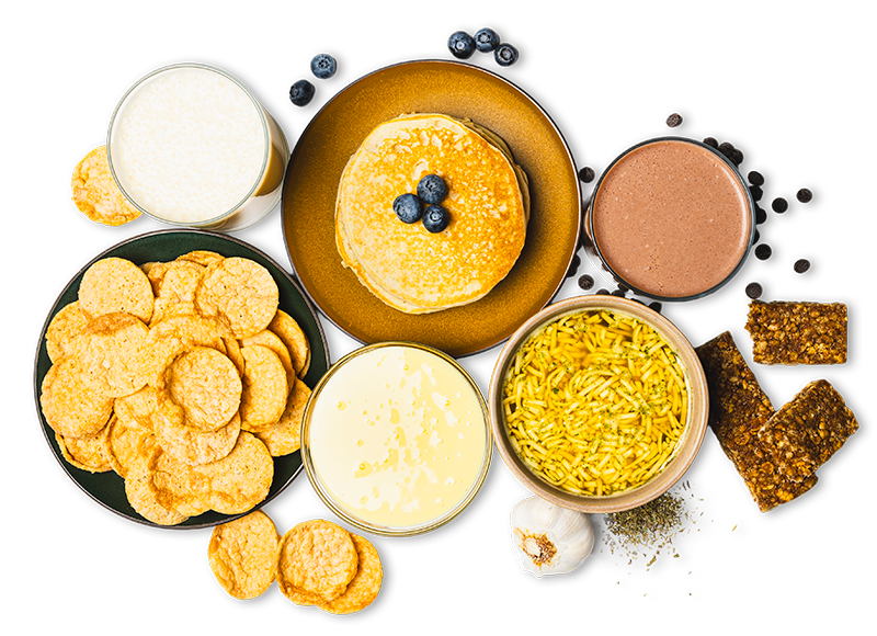 Top view of assorted snacks and drinks including pancakes with blueberries, protein chips, protein shakes, a bowl of protein soup, and protein bars