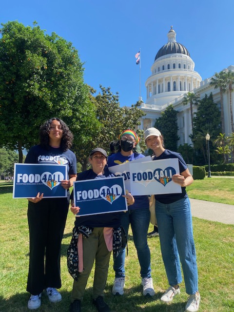 Four advocates stand proudly in front of the California State Capitol holding “Food4All” signs, symbolizing their commitment to equitable access to food and a just future for all communities.