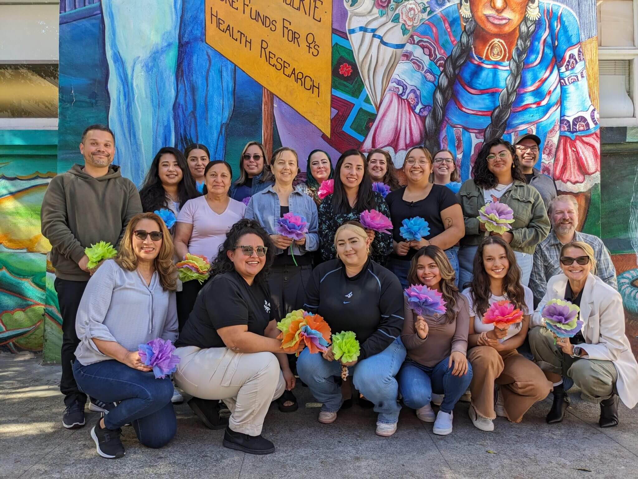 A diverse and smiling group of staff members from The Women's Building pose in front of a vibrant mural, holding colorful paper flowers, symbolizing unity, community, and empowerment.