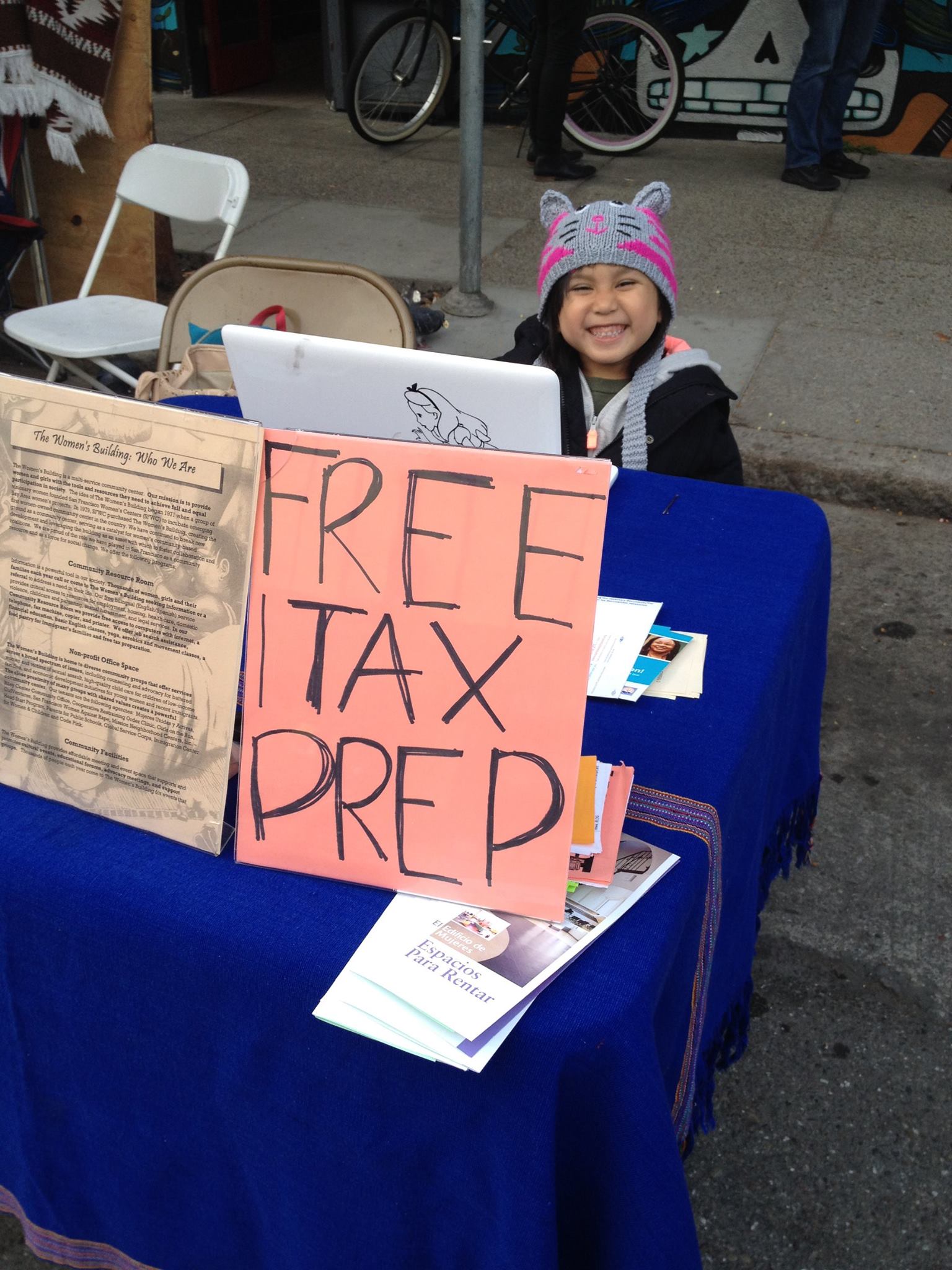 Child in a pink and gray cat hat smiling behind a table offering free tax preparation services.
