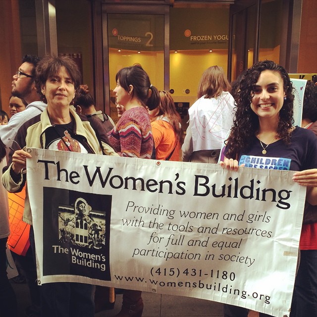 Two women hold a large banner for The Women's Building during a community rally, with people gathered in the background.