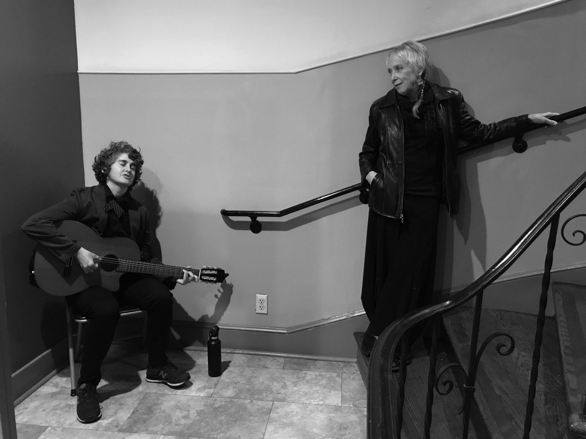 A woman plays guitar while seated at the base of the stairs as an older woman in a leather jacket looks on during a performance inside The Women’s Building.