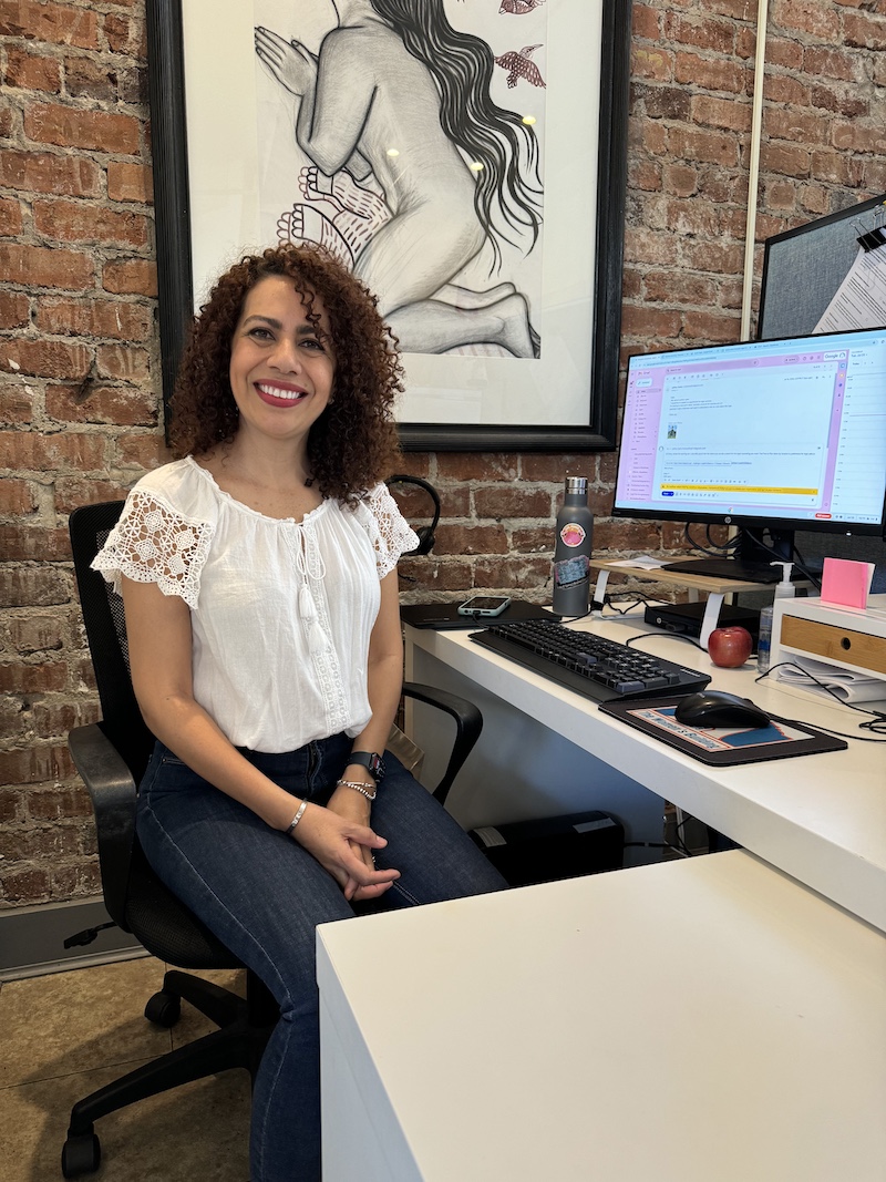 A smiling woman seated at a desk in an exposed brick office, ready to assist with housing and referral services.
