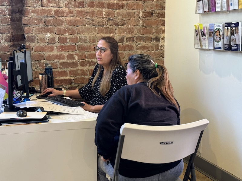 A staff member at a computer assisting a community member in an intake session at a resource clinic.