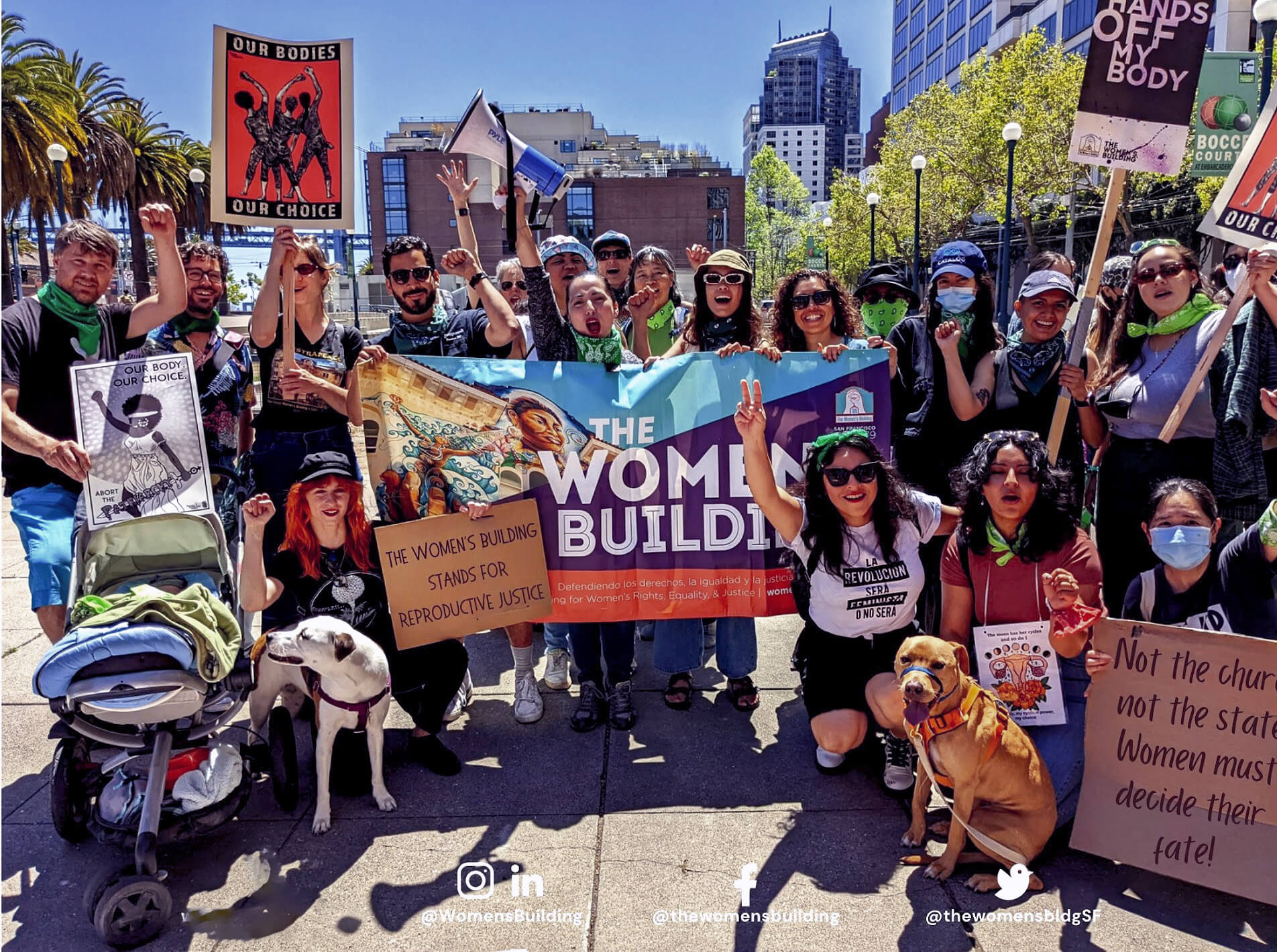 A large group of staff and community members and activists rally outdoors holding signs and a banner for The Women’s Building, advocating for reproductive justice and women’s rights.