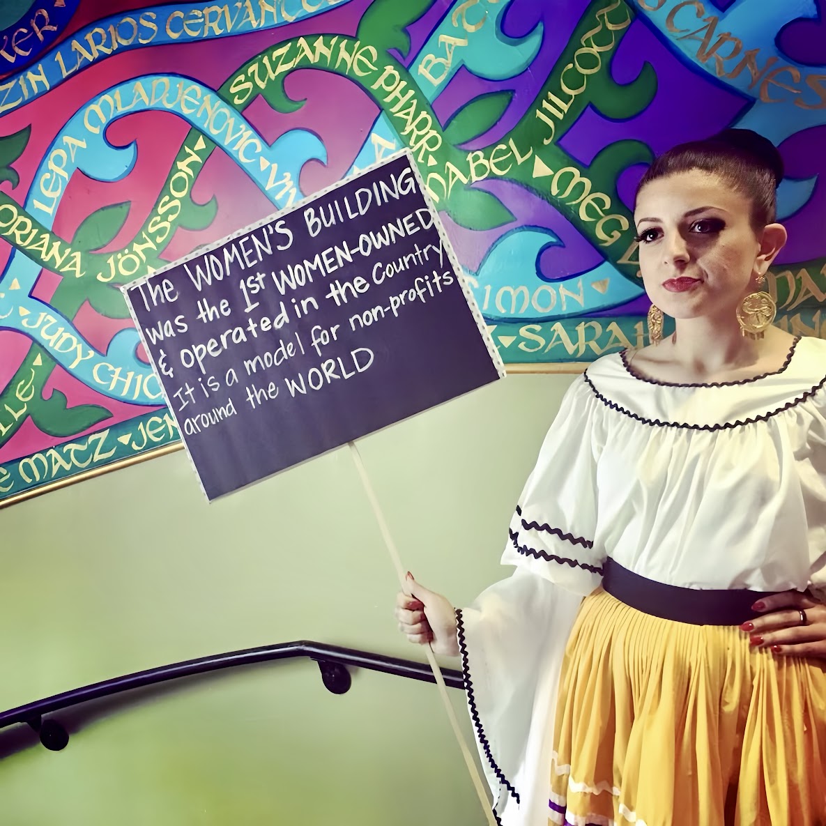 A woman in traditional dress holds a sign that reads, “The Women’s Building was the 1st women-owned & operated in the country. It is a model for non-profits around the world,” standing in front of a mural honoring feminist leaders.
