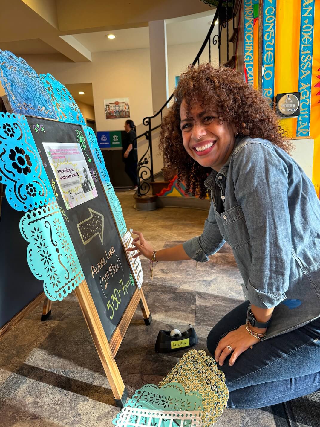 A smiling woman decorates a signboard with colorful papel picado for an event at The Women’s Building, embodying the warmth and vibrancy of community-centered work.