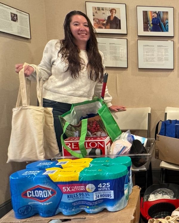 A woman holds tote bags filled with essential supplies, standing in a room with donation items at The Women’s Building.