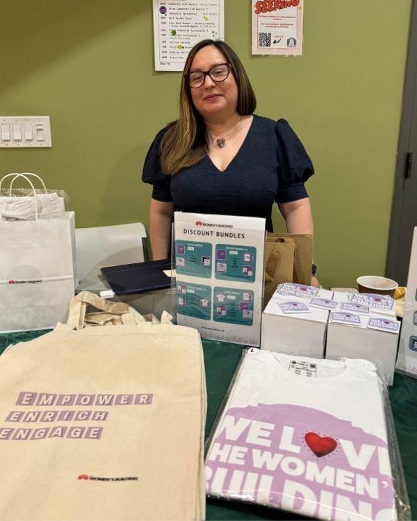 A volunteer stands behind a merchandise table featuring The Women’s Building tote bags and t-shirts to raise funds and awareness.