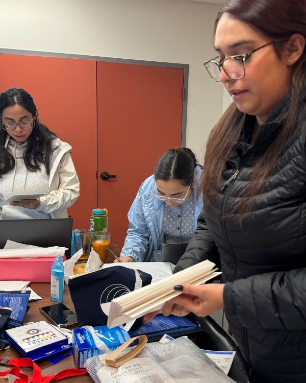 Three women from The Women’s Building staff preparing informational packets and supplies at a table filled with materials and drinks.