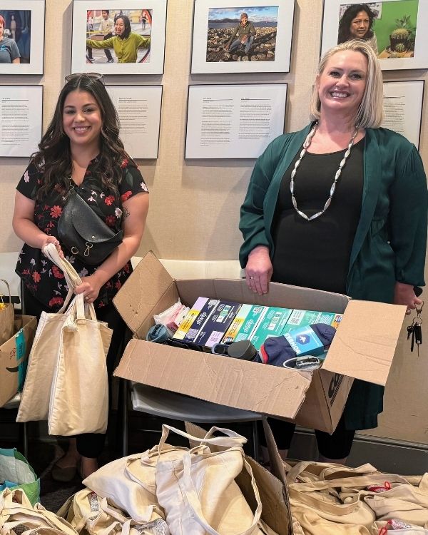 Two smiling women from The Women’s Building stand with a large box of donated items and tote bags ready for community distribution.