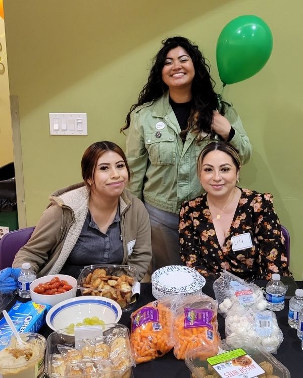 Three smiling women, including two seated at a table with snacks and one standing behind with a green balloon, during a community gathering at The Women’s Building.