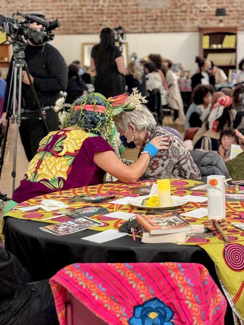 Two women share an intimate, respectful moment during a community event—one wearing a vibrant floral headdress, the other gently leaning in—surrounded by colorful table decor and books, symbolizing cultural strength and solidarity.