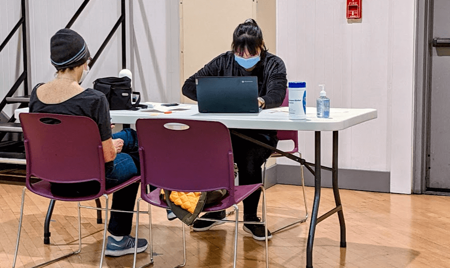A masked staff prepares a tax return for a community member seated across the table, with sanitizer and disinfectant wipes placed between them.