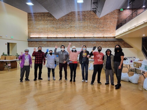 A group of masked volunteers stand together and wave inside a large hall, surrounded by food donation supplies, ready to support the community through the food pantry and CalFresh outreach.
