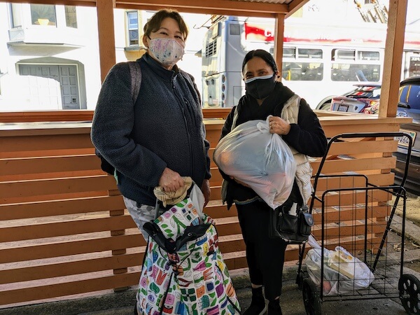 Two women smile behind their masks after receiving groceries, standing beside a shopping cart filled with food items provided through the CalFresh benefits and community food pantry support.