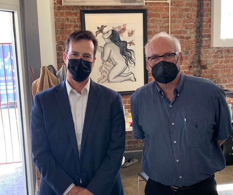 Two volunteer attorneys wearing masks pose at The Women’s Building legal clinic, standing in front of a framed artwork and exposed brick wall.