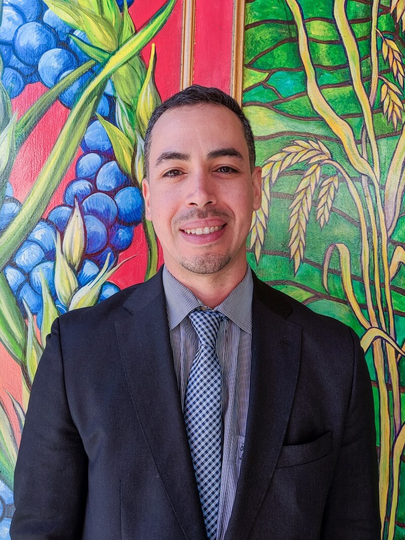 Javier Rivera, an immigration attorney, stands smiling in front of a colorful mural at The Women’s Building.