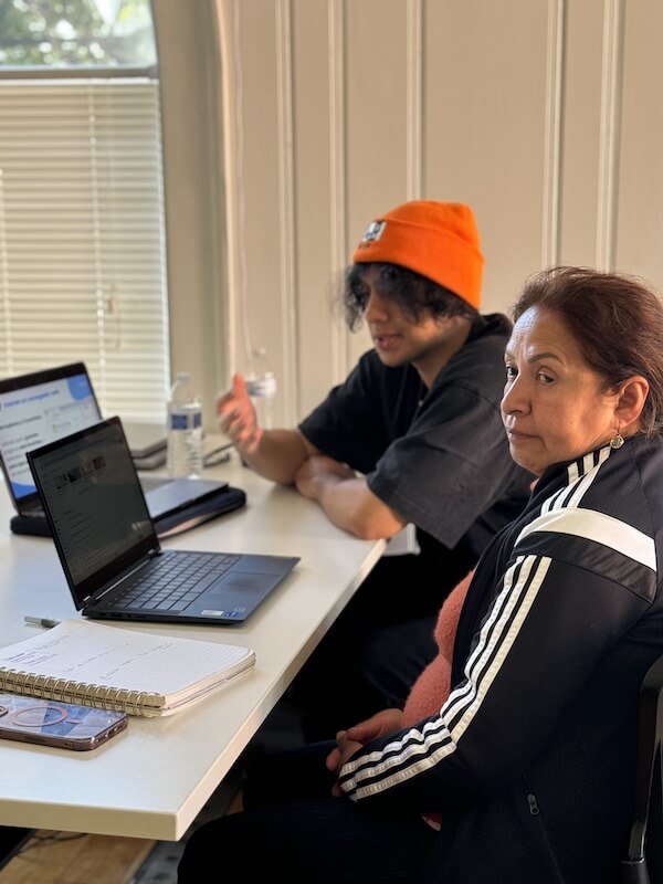 A young volunteer member wearing an orange beanie provides computer help to a community member at The Women’s Building’s Mobile Lab, with laptops and notebooks on the table.