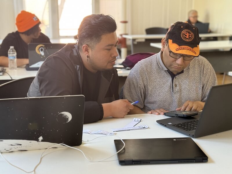 A volunteer member assists a participant with navigating a laptop and paperwork at The Women’s Building’s Mobile Lab, as others work in the background.