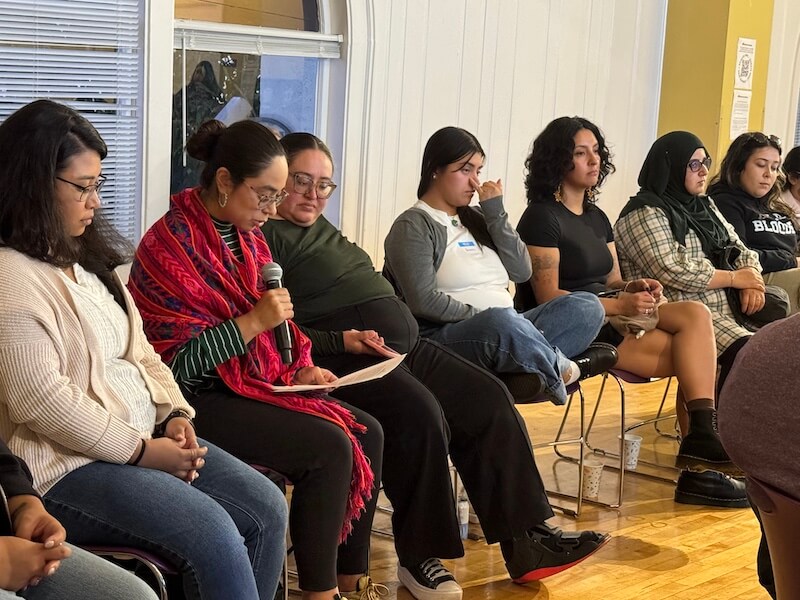 A group of young women sit in a circle, attentively listening as one person reads aloud into a microphone during a community support session.
