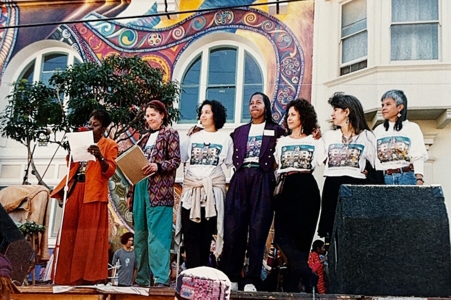 Muralists standing together on stage in front of The Women’s Building mural, wearing matching commemorative shirts during the mural public celebration.