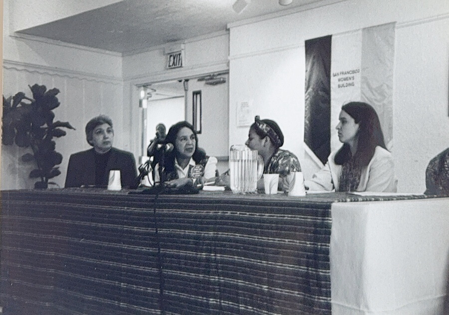 A black-and-white photo of four Latina leaders seated at a long table with microphones during a panel discussion at The Women’s Building.