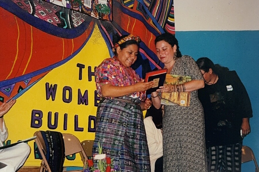 An Indigenous woman in traditional dress receives a framed award during a ceremony at The Women’s Building, with the mural in the background.