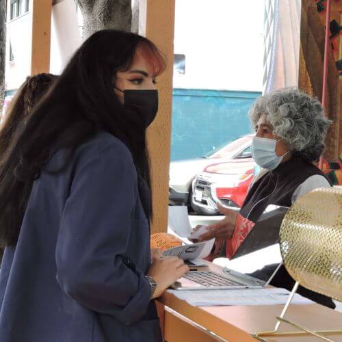 A staff member wearing a face mask assists an older woman at a check-in table during a community food distribution event.