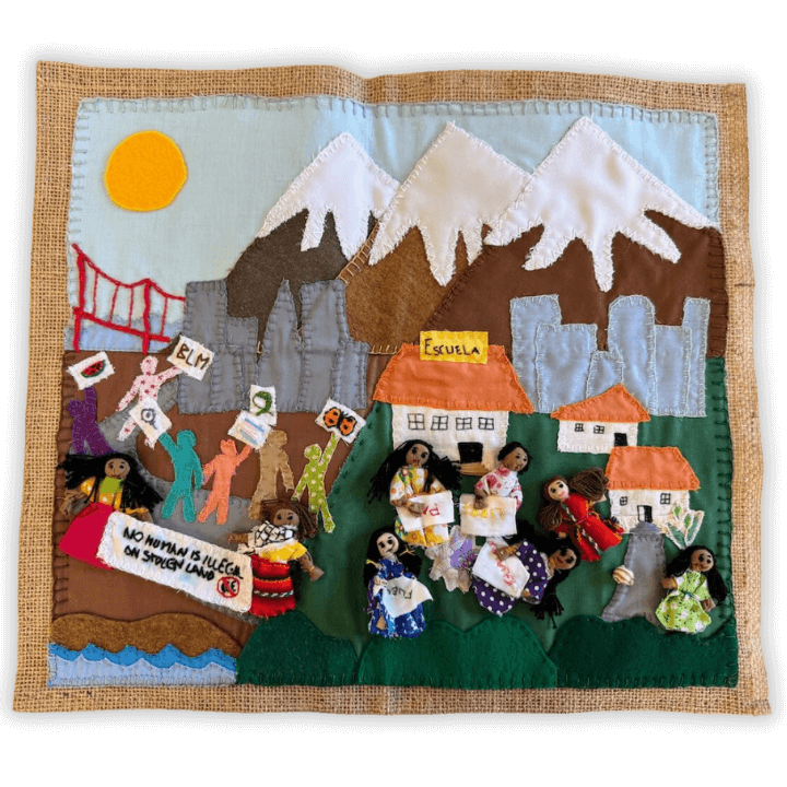 Handcrafted fabric mural showing a community scene with mountains, homes, and protest imagery.