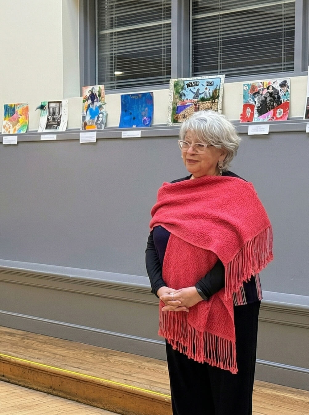Teresa Iñiguez stands in a gallery space offering an opening blessing, wearing a red shawl with artwork displayed behind her