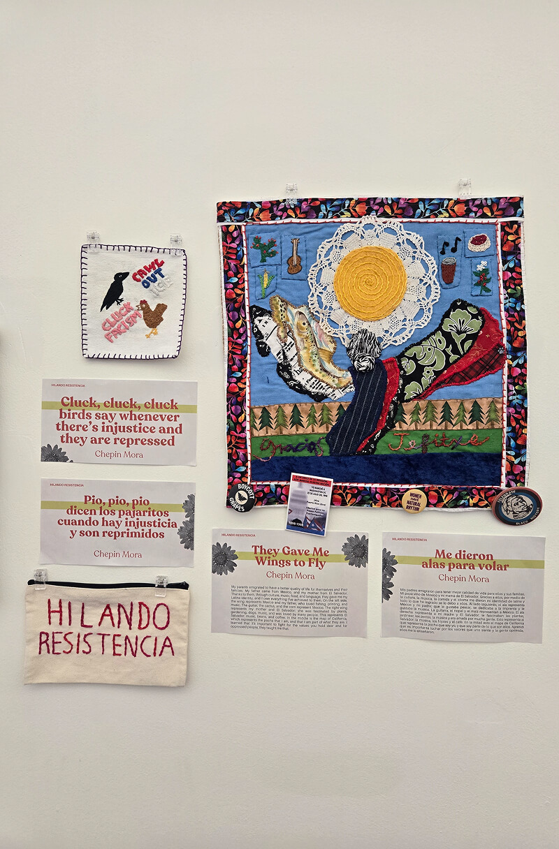 Community embroidery artworks and arpilleras displayed on a wall with stitched messages, textiles, and storytelling pieces