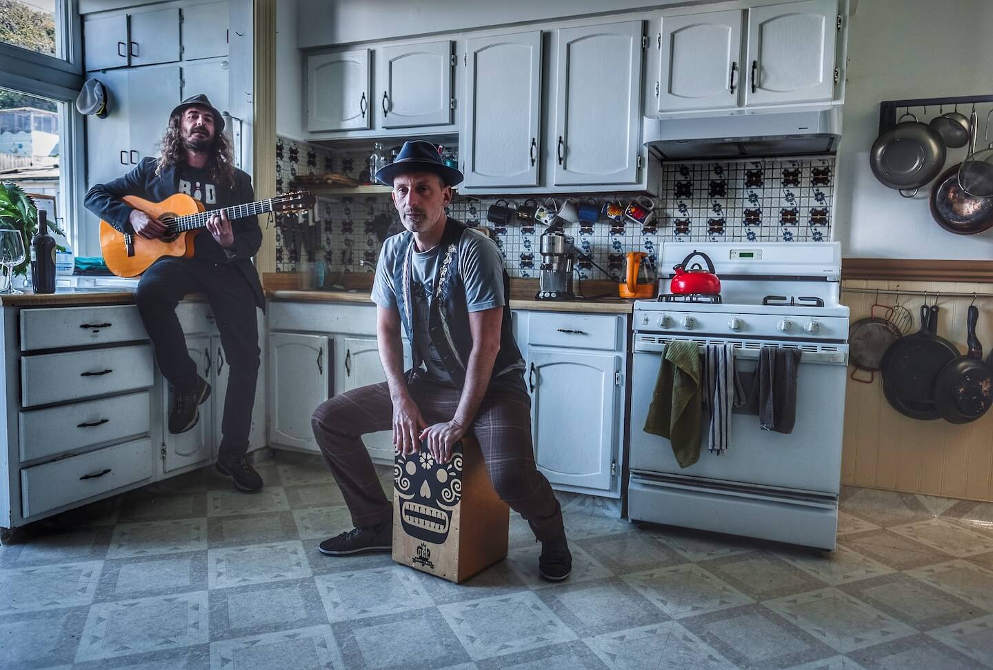 Two musicians perform Spanish and Latin music in a cozy indoor setting, one playing guitar and the other seated on a cajón drum