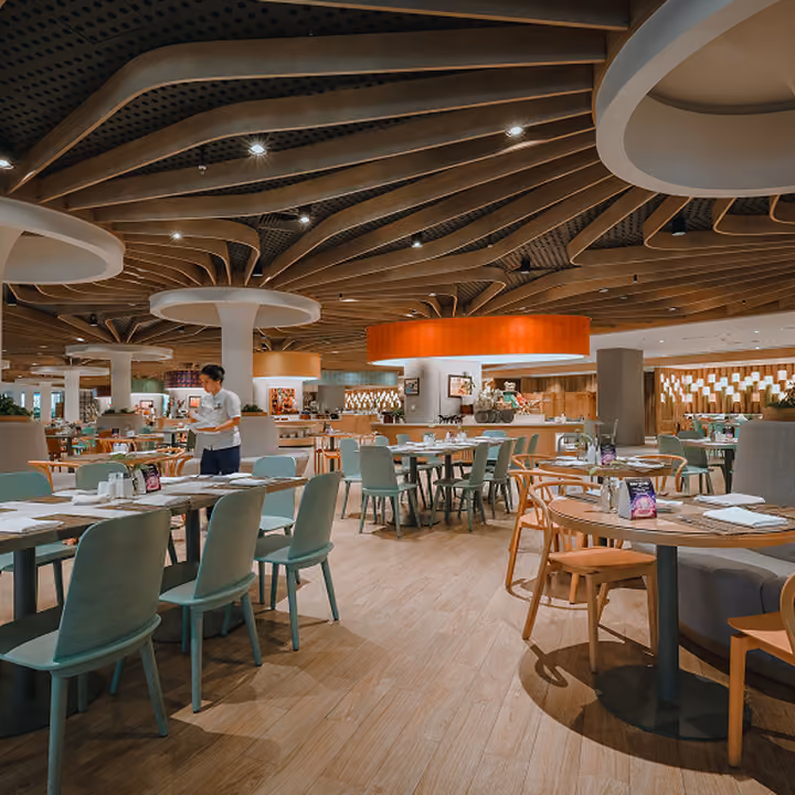 Spacious modern restaurant interior with wooden flooring, multiple tables and chairs arranged neatly, soft lighting, and a staff member attending to a table.
