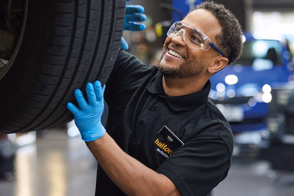 Smiling Halfords mechanic wearing safety glasses and blue gloves lifts a car tire in an auto repair shop with a blurred blue car in the background.