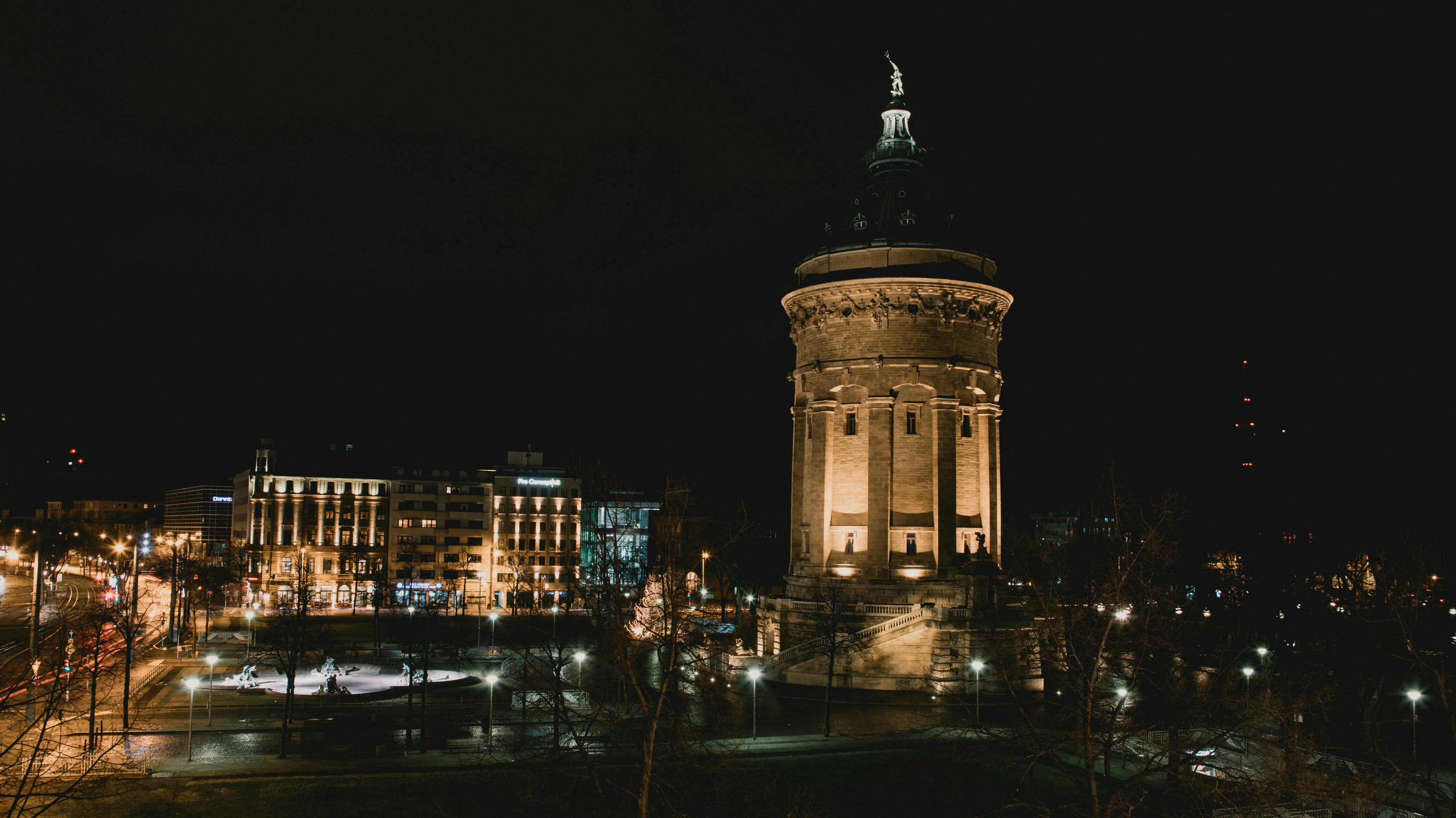 Nächtliche Stadtansicht mit einem beleuchteten Wasserturm im Vordergrund, umgeben von Straßenlaternen, Bäumen und historischen Gebäuden; im Hintergrund sind weitere Gebäude und Lichter der Stadt zu sehen.
