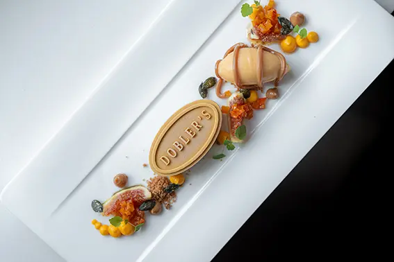 Kunstvoll angerichtetes Dessert auf einem weißen rechteckigen Teller, mit einem geformten Schokoladenelement mit der Aufschrift „Dobler’s“, einer glasierten Süßspeise, Fruchtgel-Punkten, Nüssen und dekorativen Garnierungen.

