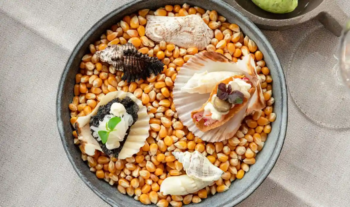 Zwei kunstvoll angerichtete Meeresfrüchte-Häppchen in Jakobsmuschelschalen, serviert auf einem Bett aus Maiskörnern, dekoriert mit Cremes, Kräutern und kleinen Muscheln in einer Schale.