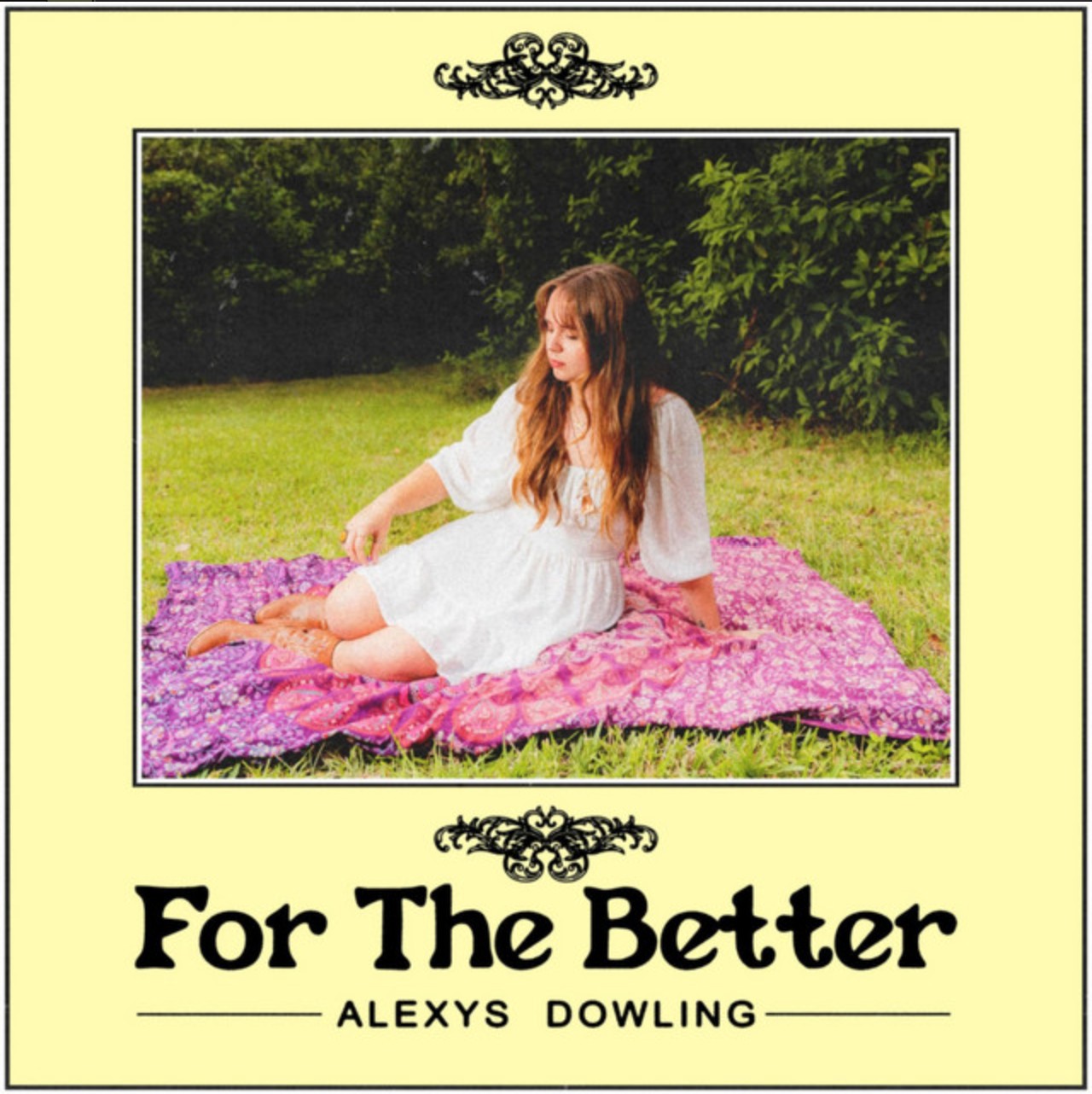 Young woman in a white dress sitting on a purple patterned blanket in a grassy area with trees behind.
