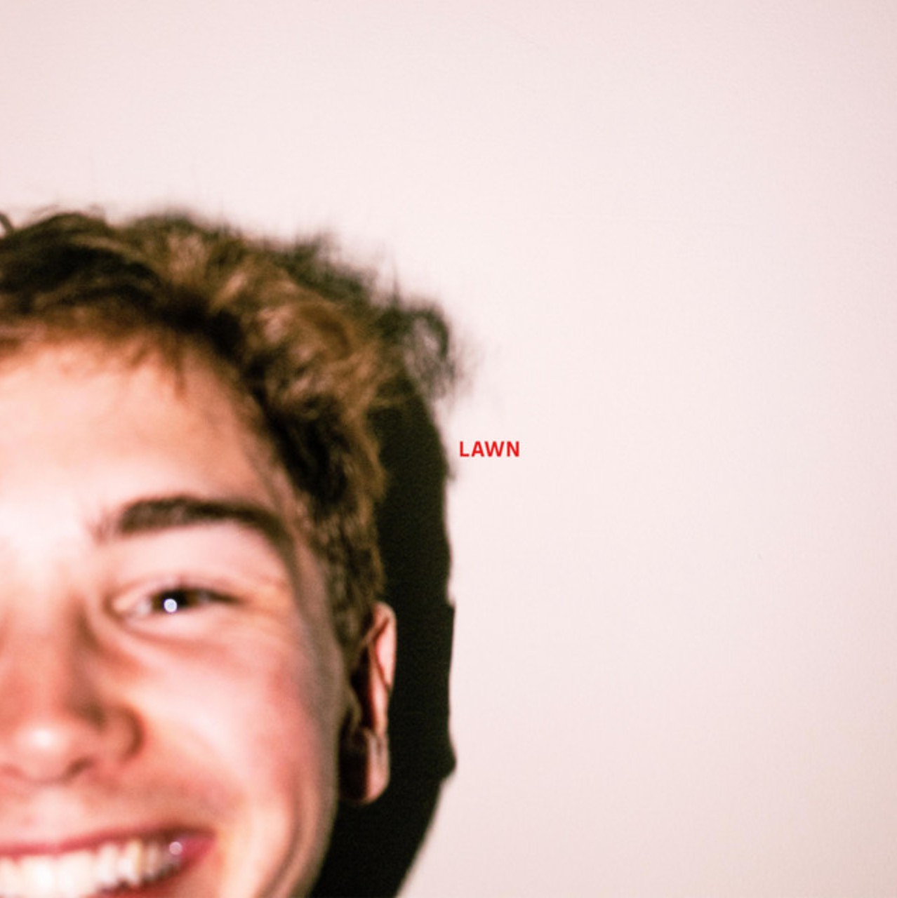 Close-up of a smiling young man with curly hair against a pale wall with the word 'LAWN' in red text.