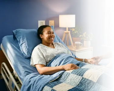 Smiling woman lying in a hospital bed holding someone's hand in a cozy room with a bedside lamp and plant.