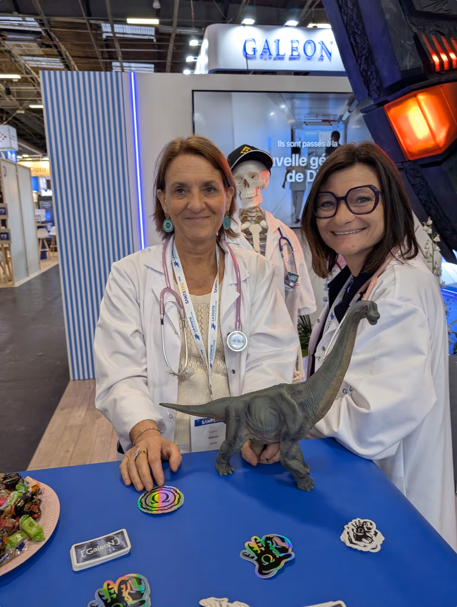 Deux femmes en blouse blanche avec stéthoscopes au stand Galeon lors du salon SantExpo, avec un dinosaure jouet