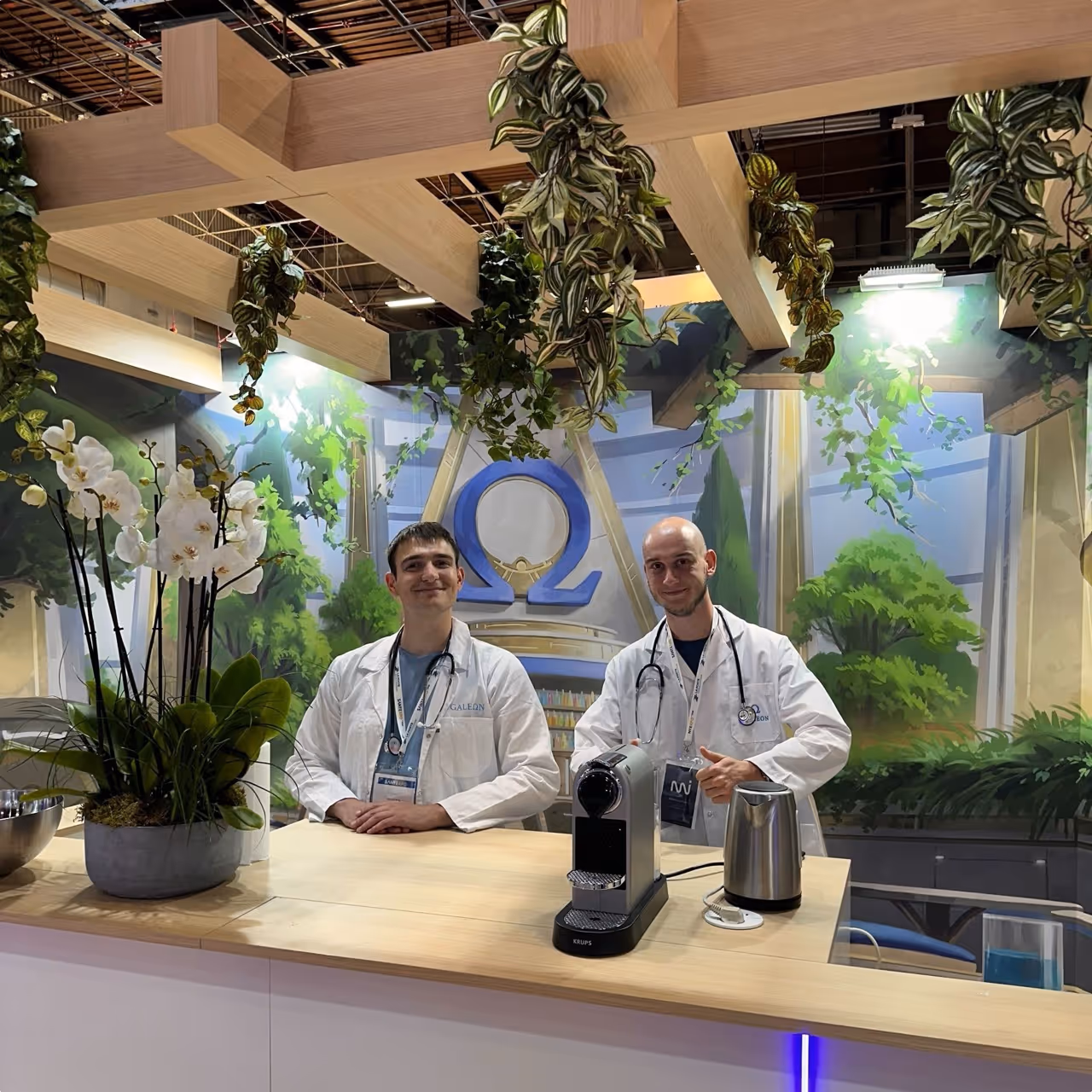 Deux membres de l'équipe Galeon en blouse blanche sur un stand d'exposition décoré de plantes et du logo Omega